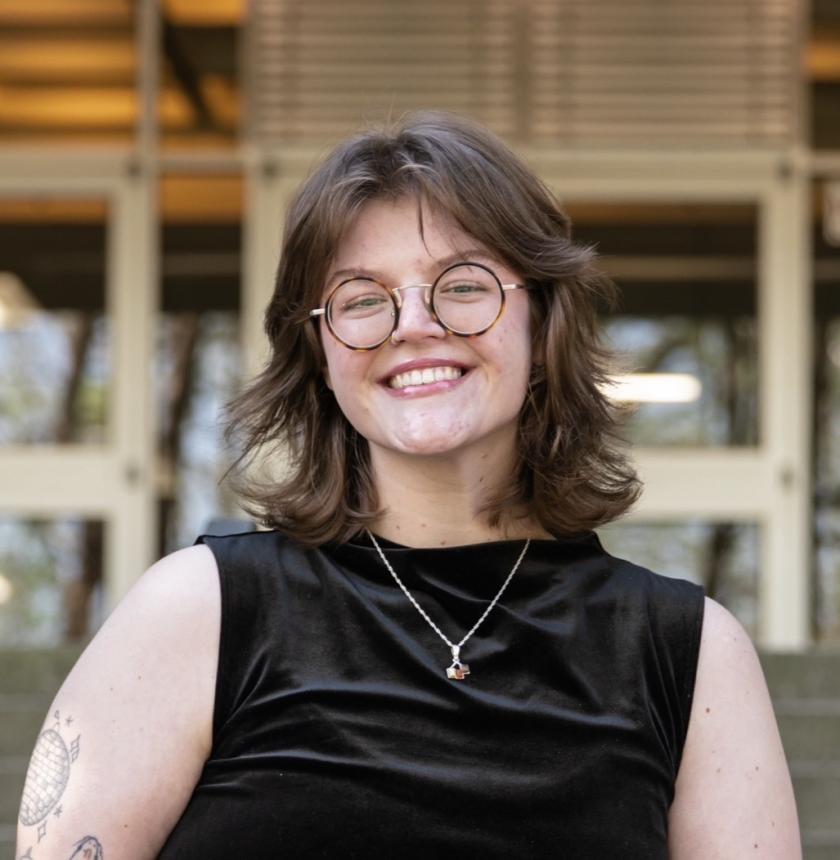 white young woman with brown hair and glasses wearing a black shirt standing in front of a building that is out of focus in the background.