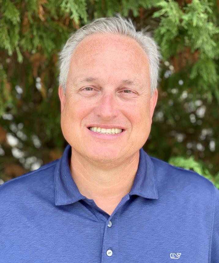 white older man wearing a dark vibrant blue shirt with green foliage in the background. 